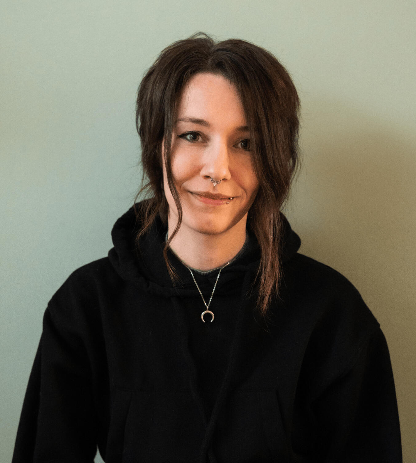 Portrait by Bambini Fotografie Author portrait of a white person with dark brown hair, cat-eye makeup, a nose and lip piercing, wearing a black sweater.